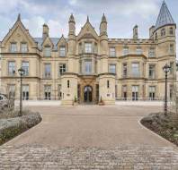 Historic stone building with pointed towers, ornate façade, and symmetrical windows, situated between two lampposts. A driveway leads to the entrance.