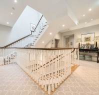 A well-lit, modern staircase with white railings and carpeted floors. There are lamps, art, and a table against the wall, adding decorative elements to the space.