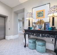 Elevator lobby with a decorative console table, lamps, framed artwork, a floral arrangement, and two cushioned stools on a patterned carpeted floor.