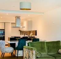 Modern open-plan living area with green sofa, patterned chairs, and a white dining table. Kitchen features stainless steel appliances and black accents. Minimalist decor with a large wall frame.