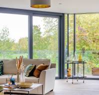 Bright living room with large windows, a beige sofa with colorful pillows, a glass and metal side table, and an armchair. Greenery and trees visible outside.
