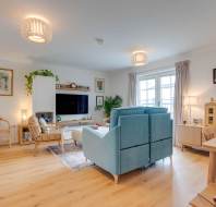 A cozy living room with light wood flooring, a blue sofa, beige chairs, a TV on a white wall unit, potted plants, and a small wooden dollhouse. Natural light enters through the window.