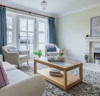 Modern living room with a white sofa, two armchairs, glass coffee table, and fireplace. Blue curtains frame large windows. A gray rug and décor, including plants and artwork, complete the scene.