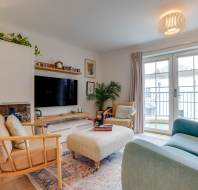 Bright living room with a blue sofa, two wooden chairs, a cushioned ottoman, TV, plants, and a balcony view through large glass doors. Chandelier-style light fixture and decorative elements present.