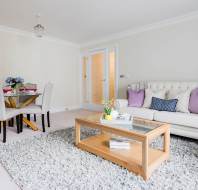 Tastefully decorated living room with a white sofa, purple cushions, a wooden coffee table, and a dining area with a round table and four chairs on a gray carpet.