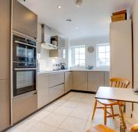 Modern kitchen with gray cabinets, double ovens, and small dining table with two wooden chairs, next to a vase of flowers. Light-colored tiled floor and two windows in the background.
