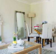 A dining area with a table and chairs, a large mirror, and a vase of flowers. A teapot and cup sit on a tray on the coffee table. Neutral tones with a light, airy atmosphere.