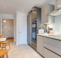 Modern kitchen with beige cabinetry, built-in oven, white countertops, and tile flooring. A small wooden dining set is in the corner, and a hallway is visible in the background.