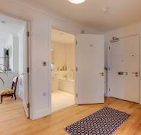 A hallway with wooden flooring leads to an open bathroom door. A patterned rug is on the floor, and a potted plant is on a glass table. A chair is positioned in the living area.