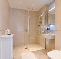 Modern bathroom with a glass-enclosed shower, beige tiles, wall-mounted sink, toilet, chrome towel rack, and a small white cabinet with toiletries.