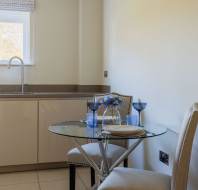 A bright kitchen with a round glass table, two beige chairs, and tabletop items. There's a window and a sink with modern cabinets underneath.