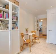 A small dining area in a kitchen with two wooden chairs, a table, and a cabinet filled with books and kitchen items. The floor is tiled, and there are overhead lights.