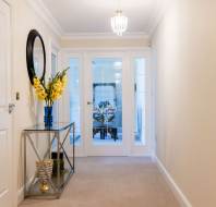 Hallway with a glass table and flowers on the left, leading to a glass door showing a dining room set. White walls and a ceiling light fixture are visible.
