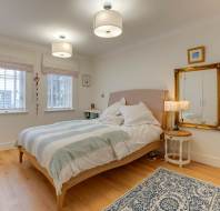 A cozy bedroom with a neatly made bed, two windows with blinds, a rug on wooden flooring, a bedside table with a lamp, and a mirror on the wall.