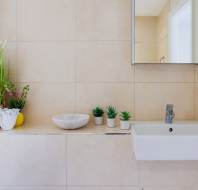 Bathroom with beige tiles, a white sink, a mirror, plants in pots, a soap dispenser, and decorative items including a woven basket on the counter.