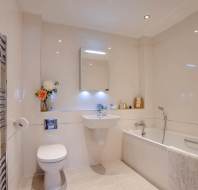 Modern bathroom with a toilet, wall-mounted sink, bathtub with a towel, mirror cabinet, flowers in a vase, and towel radiator. Beige tiles cover the walls and floor.