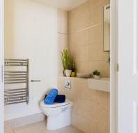 Modern bathroom with beige tiled walls, featuring a white toilet, towel radiator, and sink. A blue towel is placed on the toilet, and two plants adorn the shelf above.