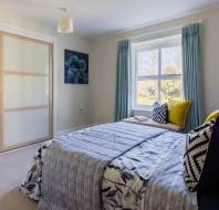 A neatly arranged bedroom with a bed featuring patterned bedding, pillows, and a throw. There's a window with blue curtains, sliding closet doors, and a framed picture on the wall.