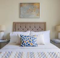 A tidy bedroom with a bed featuring white and patterned pillows, flanked by glass-topped nightstands and white lamps. A textured abstract painting hangs above the tufted headboard.