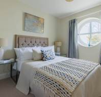 A neatly arranged bedroom with a bed, pillows, a patterned blanket, bedside lamps, and a large round window with blue curtains. A decorative painting hangs above the bed.