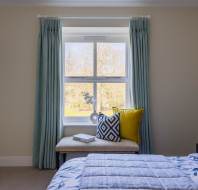 A bedroom with a window featuring light blue curtains, a bench with decorative pillows, a bed with a floral duvet, and a lamp on a nightstand. A picture frame hangs on the wall.