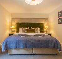 A tidy bedroom with a large bed featuring a green upholstered headboard, two wall-mounted lamps, a beige carpet, and framed pictures on the wall.