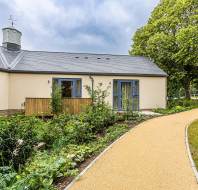 Modern, single-story house with beige walls, blue-framed windows, and a slate roof. A curved gravel path leads to the entrance, surrounded by greenery and trees under a cloudy sky.