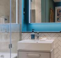 Modern bathroom with a glass shower, white sink with a blue soap dispenser, and a large mirror. The walls are painted teal and tiled with a chevron pattern.