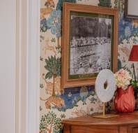 Framed black and white photo on a wallpapered wall with nature and animal patterns, beside a wooden table with flowers, a lamp, and a decorative object. Kitchen visible through an open door.