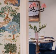 A dining area with a round table set for a meal, four blue chairs, and a vase with leaves. A wall with a colorful nature-themed wallpaper is partially visible.