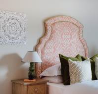 Bedroom with a decorative pink patterned headboard, two green pillows, and a wooden bedside table with a lamp. Wall art with an intricate white pattern hangs above the bed.