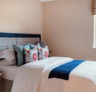 A neatly made bed with patterned pillows and a blue throw, next to a wooden side table with a lamp, in a room with beige walls and a window with a Roman shade.