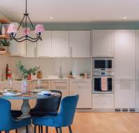 Modern kitchen featuring white cabinets, built-in ovens, a round dining table with blue chairs, and a black chandelier with pink shades. A large wall clock is visible.