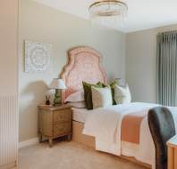 A neatly arranged bedroom featuring a bed with orange patterned headboard, green and white pillows, bedside table with lamps, and a window with drapes. A decorative wall piece hangs above.