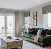A cozy living room with a green sofa, patterned pillows, a floral ottoman, and a blue chair. Large windows with curtains allow natural light. Wall art and decor complete the space.