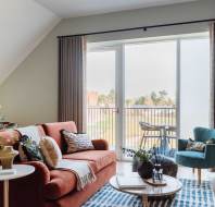 Modern living room with a red sofa, teal armchair, and coffee table on a patterned rug. Large windows open to a balcony with a table and chairs. Dining area visible in the background.