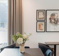 Dining area with a black table, navy chairs, and a white vase of flowers. A modern chandelier hangs above. Framed artwork is displayed on a wall with curtains beside a window.