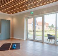 Spacious room with wooden flooring, two exercise mats, a blue kettlebell, and a kitchenette. Large glass doors open to a garden view.