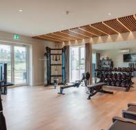 A modern gym with exercise machines, weights, and workout benches. Large windows and a mirrored wall enhance the spacious interior.
