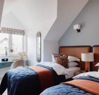 A cozy bedroom features two twin beds with blue quilts and orange throws, a blue desk by the window, and a mirror on the wall. A lamp and decorative pillows complete the space.