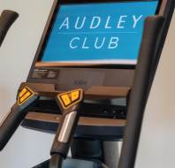 Close-up of a stationary bike display screen showing the text "AUDLEY CLUB" with handlebars in the foreground.