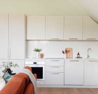 Modern kitchen with white cabinets, built-in oven, and countertop accessories. A brown sofa with cushions partially visible in the foreground. Light wood flooring and a plant accent.