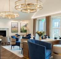 A modern lounge with blue velvet chairs, circular chandeliers, and a fireplace. Large windows, a wooden coffee table with flowers, and framed artwork decorate the space.