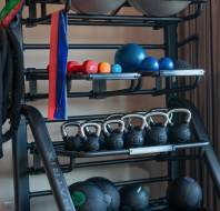 A gym rack with exercise balls, resistance bands, colorful dumbbells, kettlebells, medicine balls, and a foam roller arranged neatly on shelves.