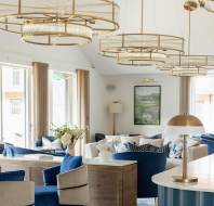 Spacious modern lounge with large windows, blue and white seating, gold lighting fixtures, and a central table with flowers.