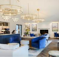Elegant hotel lounge with blue and white sofas, marble tables, and modern chandeliers. A bar with coffee machines is in the corner. Large artworks and a fireplace adorn the walls.