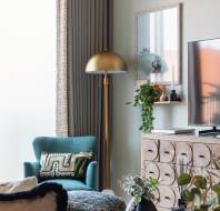 Modern living room with a green armchair, patterned cushion, and a tall floor lamp. A large TV sits on a wooden cabinet with circular designs, and a framed picture hangs on the wall.