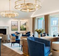 Luxurious lounge area with blue velvet chairs, round chandeliers, large windows, and framed artwork. Neutral tones with gold accents create an elegant atmosphere.