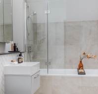 Modern bathroom with a white toilet, floating vanity, and bathtub with glass shower screen. Beige tiles, a large mirror, and a skylight enhance brightness. Dried flowers on a bathtub tray.