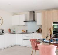 Modern kitchen with white cabinets, light wood accents, and pink velvet chairs. A dining table is set with dishes, and a large wall clock is visible. Green plants add a touch of color.